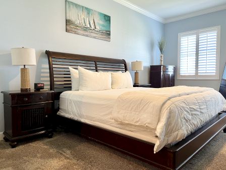 A bedroom with a wooden bed, white bedding, bedside tables, lamps, a painting above the bed, and a window with white shutters.