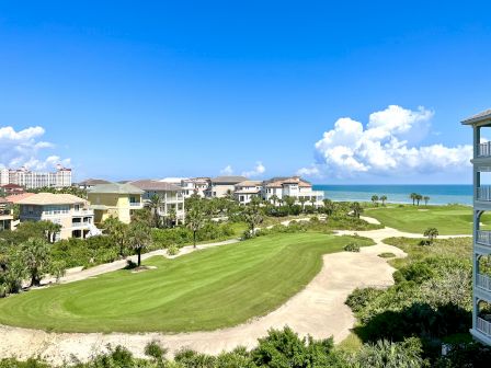 Coastal golf course view with green, sand traps, palm trees, nearby houses, blue ocean, and a clear sky with fluffy clouds in the distance.