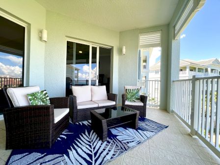 Outdoor patio with wicker furniture, green leaf pillows, a glass-top table, and a leaf-patterned rug, overlooking neighboring buildings.