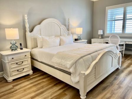 A bedroom with a white bed, nightstands with lamps, and a desk with a chair and window shutters.