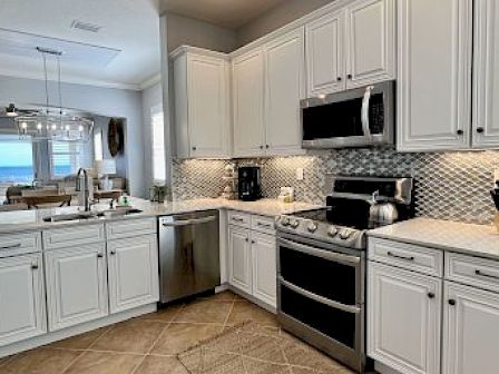 A modern kitchen with white cabinets, stainless steel appliances, and a tiled backsplash. There's an ocean view from the adjacent room.