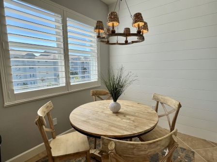 A dining area with a wooden table, chairs, a plant centerpiece, and a woven light fixture. Large windows show a view outside.