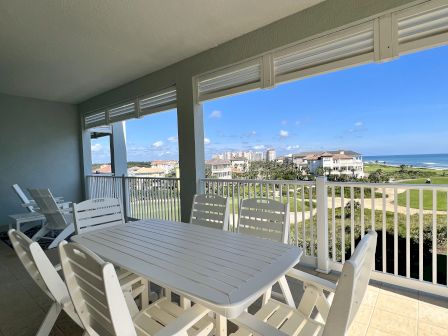 A covered outdoor patio with white furniture, offering views of a golf course, houses, and ocean under a clear blue sky.