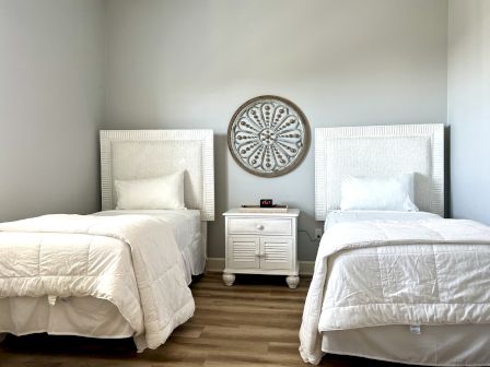 The image shows a bedroom with two twin beds, a white nightstand, and a decorative wall piece, all set against a light gray wall.