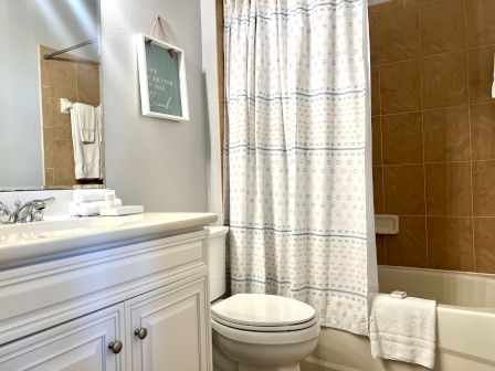 A bathroom with a white vanity, stacked towels, decorative sign, toilet, and bathtub with a curtain.