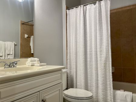 A bathroom with a sink, mirror, and countertop with towels and toiletries; nearby, a shower/tub with a white curtain and tan tiles.