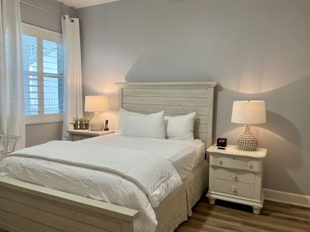 A bedroom with a bed, wooden headboard, two nightstands, lamps, and a digital clock. White curtains cover a window to the left.
