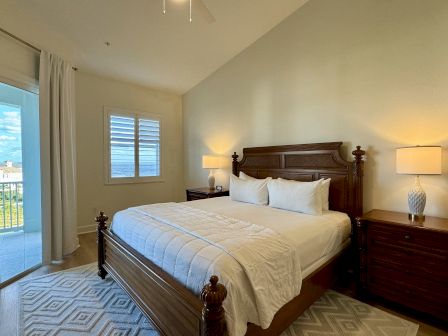 A cozy bedroom with a large bed, wooden headboard, white bedding, and nightstands with lamps. There's a window and a balcony door.