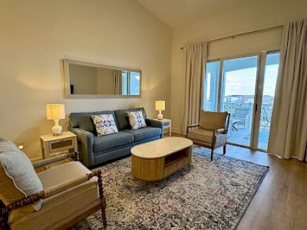 A cozy living room with a sofa, armchairs, a coffee table, and lamps, overlooking a balcony with a view through glass doors.