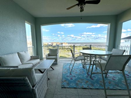 A cozy balcony with a ceiling fan, outdoor seating, a round table, and a colorful rug overlooks a scenic view of houses and the ocean.