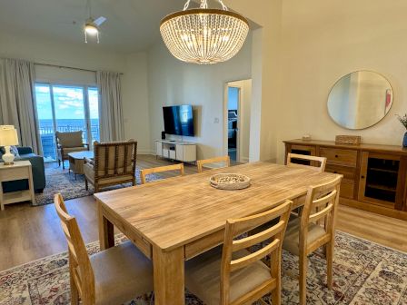 A cozy living-dining area features wooden furniture, a TV, chandelier, and a large window with ocean view.