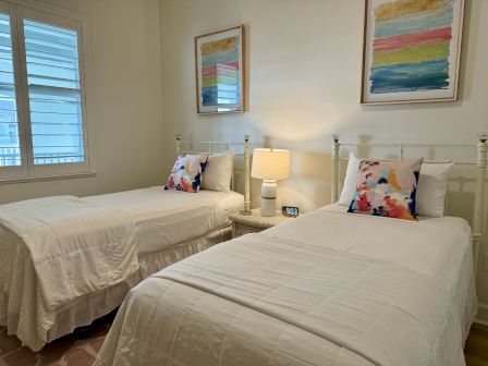 A bedroom with two white twin beds, colorful pillows, abstract art, a bedside table, lamp, and a clock reading 9:03.