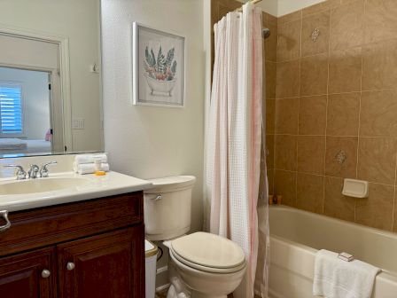 The image shows a bathroom with a sink, a toilet, and a bathtub with a shower curtain. Towels and toiletries are on the vanity.