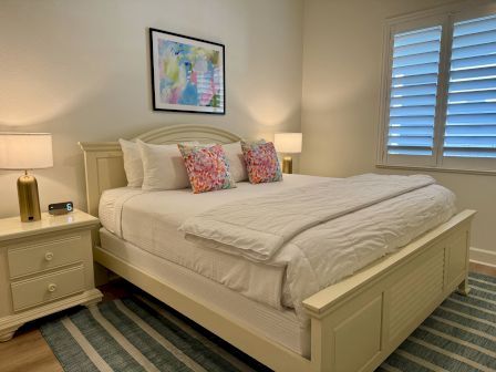 A cozy bedroom with a large bed, colorful pillows, bedside tables with lamps, abstract wall art, closed shutters, and a striped rug.