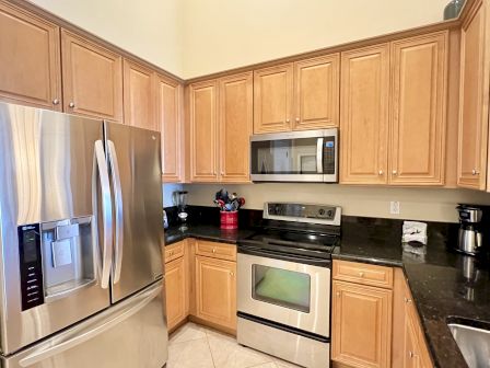 A modern kitchen with wooden cabinets, stainless steel appliances, including a fridge, stove, and microwave. Countertops hold various utensils.