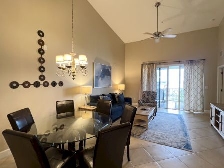 A living area featuring a glass dining table, leather chairs, a blue couch, wall art, ceiling fan, and large windows with patterned curtains.