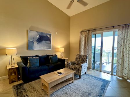 A cozy living room with a dark sofa, patterned armchair, and wooden table. Lamps flank the sofa; a painting hangs above.