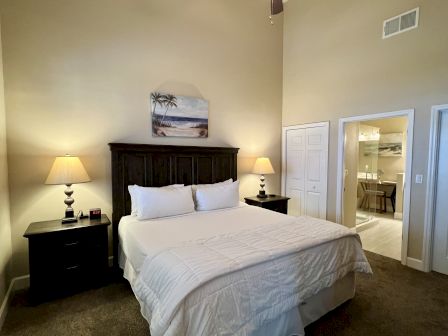 A cozy bedroom with a large bed, two bedside tables with lamps, a painting above, and a view into a bathroom.
