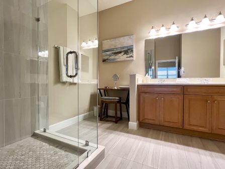 A bathroom with a glass shower, wooden cabinets, large mirror, sink, and a small vanity table with a chair.