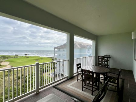 A balcony with wooden furniture overlooks a green lawn, palm trees, and the ocean. A building is visible in the distance.