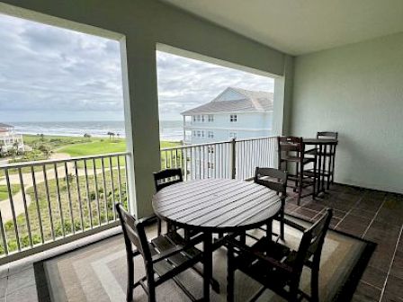 A covered balcony with tables and chairs overlooks a coastal view. The ocean, buildings, and greenery are visible.