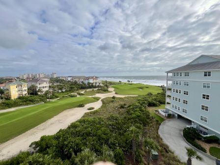 A coastal view featuring a golf course, residential buildings, and the ocean under a partly cloudy sky.