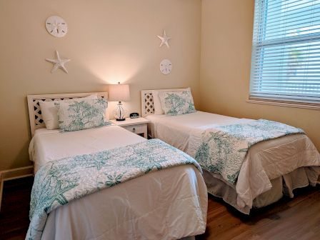 The image shows a bedroom with two twin beds, nautical-themed decor, a lamp on a shared nightstand, and a window with blinds.