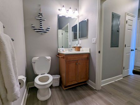 A bathroom with a toilet, vanity, mirror, and decorative anchor wall piece. Bathrobe and wall-mounted toilet paper holder are visible.