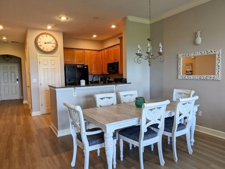 A dining area with a chandelier, table, and six chairs. A clock is on the wall, and the kitchen is visible in the background.
