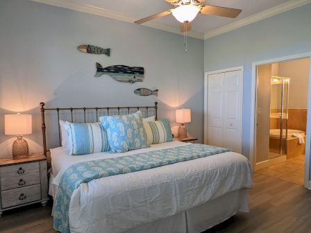 A cozy bedroom with a bed, blue and white bedding, fish wall art, two lamps on nightstands, and an open door leading to a bathroom.