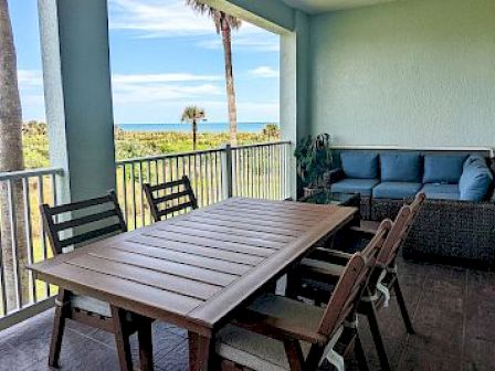 A patio with a wooden table, chairs, and a sofa overlooks a scenic ocean view with palm trees under a blue sky.