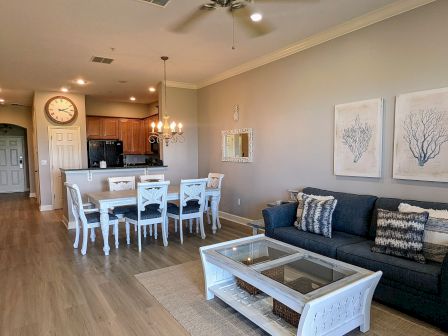 The image shows a living room and dining area with a blue sofa, white furniture, coastal decor, and a kitchen with wooden cabinets.