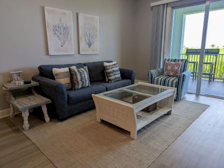 The image shows a cozy living room featuring a blue sofa, striped armchair, coffee table, wall art, and a view through sliding glass doors.