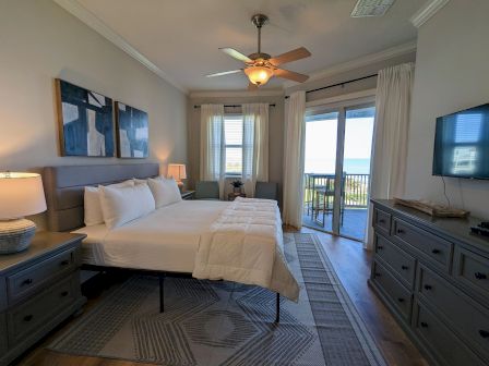 A cozy bedroom with a large bed, abstract wall art, ceiling fan, balcony view, dresser, and a TV on the wall.