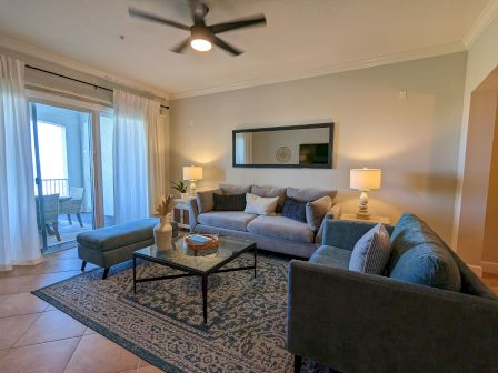 A cozy living room with sofas, a glass coffee table, lamps, a rug, and a ceiling fan, leading to a balcony with seating.