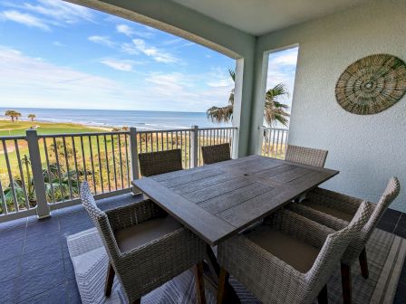 An outdoor patio with wicker chairs around a wooden table, overlooking a scenic ocean view and palm trees.