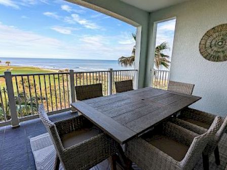 Outdoor patio with a wooden dining table, wicker chairs, ocean view, and wall decor under an open sky.