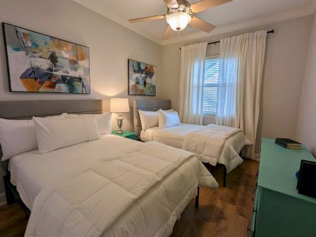 A cozy bedroom with two neatly made beds, abstract wall art, a teal dresser, a lamp, and a window with white curtains.