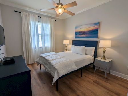 A cozy bedroom with a bed, two lamps, a ceiling fan, curtains, and a TV. A beach painting accents the wall above the bed.