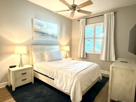A cozy bedroom with a bed, two lamps, a painting above the headboard, a window with white curtains, and a ceiling fan.