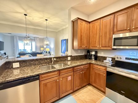 A kitchen with wooden cabinets, granite countertops, stainless steel appliances, and a view into a living area with pendant lights.