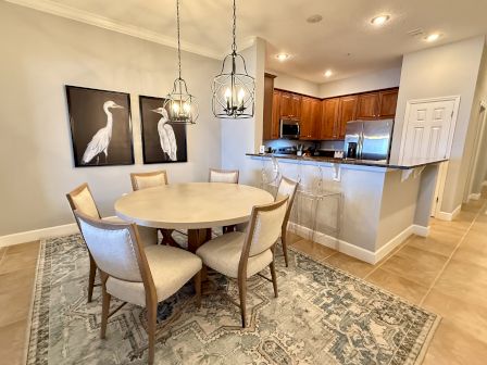 A dining area with a round table, six chairs, pendant lights, bird artwork, and a kitchen with wooden cabinets and stainless appliances.