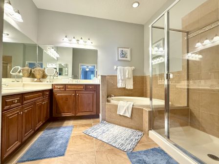 A bathroom with wooden cabinets, dual sinks, a bathtub, glass shower, mirrors, blue rugs, and warm lighting ends the sentence.