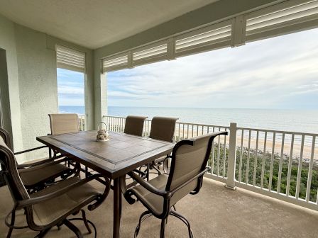 A balcony with a table and chairs overlooks the ocean, featuring a sandy beach and a railing, under a cloudy sky.