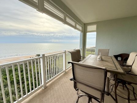 A balcony with chairs and a table overlooks the ocean, offering a serene beach view with greenery below and a cloudy sky above.