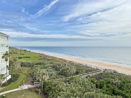 A coastal scene shows a building, a golf course, lush greenery, a sandy beach, and the ocean under a partly cloudy sky.
