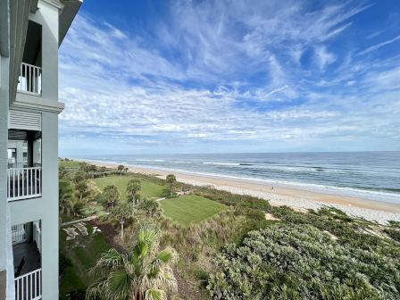 View of a beach with ocean waves, grass, palm trees, and a building with balconies on the left under a partly cloudy sky.