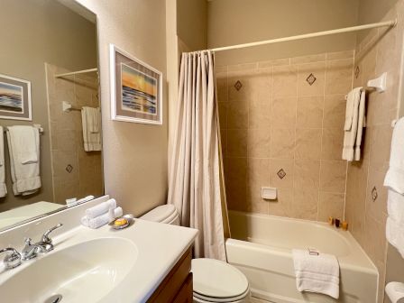 A bathroom with a sink, mirror, and bathtub with a curtain. Towels are neatly arranged on racks, and a framed picture hangs on the wall.