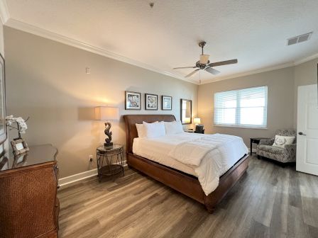 A bedroom with a large bed, nightstands, lamp, ceiling fan, framed art, dresser, and an armchair by the window.