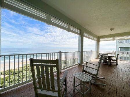 A covered patio overlooking a sandy beach and ocean, with rocking chairs and a table set, surrounded by a tiled floor and white railing.